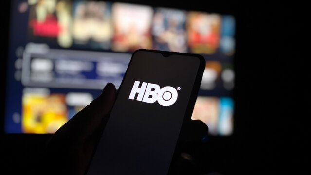Hand holding smartphone with HBO app on screen in front of TV