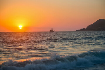 Pirate ship sailing on the Mediterranean Sea during a golden sunset in Alanya, Turkey. Beautiful coastal landscape, evening cruise, and summer vacation atmosphere at Cleopatra beach in Antalya.
