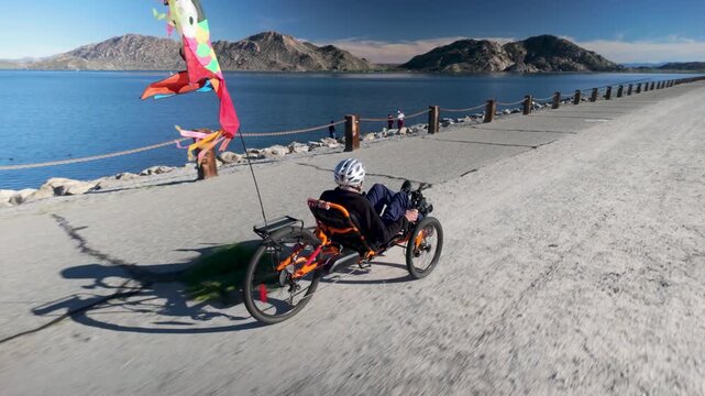 An elderly man enjoys biking on a recumbent electric trike by a lake with mountains. Colorful flags flutter in the breeze as he rides.