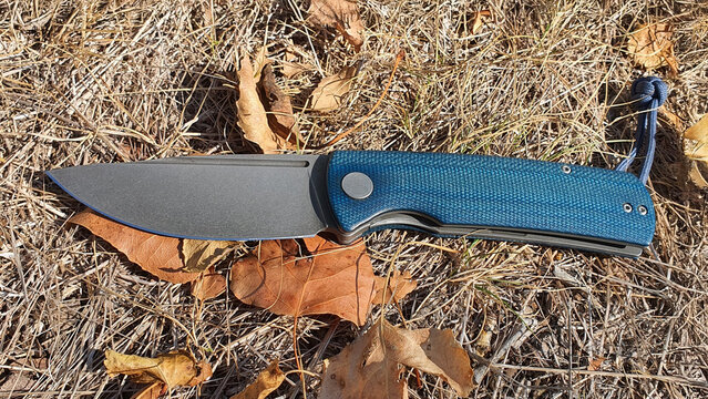 Folding pocket knife blue micarta handle stone washed blade on dry autumn grass leaves outdoor survival edc gear tactical tool professional photography