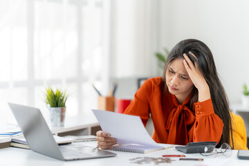 Financial Strain and Concern: A woman absorbed in deep thought, her expression etched with worry as she reviews financial documents at her desk, with a laptop, calculator, and a potted plant.