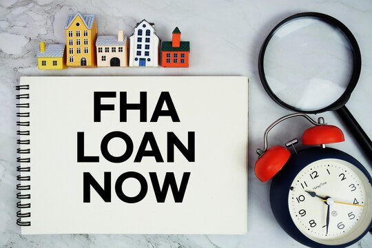FHA Loan Now text written on notepad with house model, magnifying glass and alarm clock on marble background