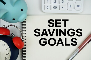 Set Savings Goals text written notepad with calculator, piggy bank and alarm clock on white background