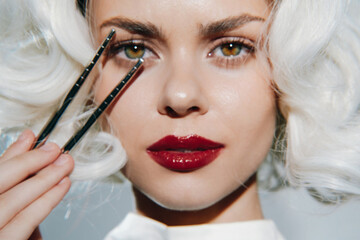 Elegant woman with vintage hairstyle applying makeup, showcasing beauty and confidence, in a soft pastel background with dramatic lighting and facial expression.