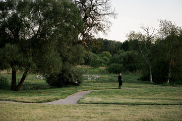 Solitary figure walking along a winding path through a lush green park at dusk, surrounded by tall trees and open meadow, evoking calm, reflection, and minimalist natural elegance.