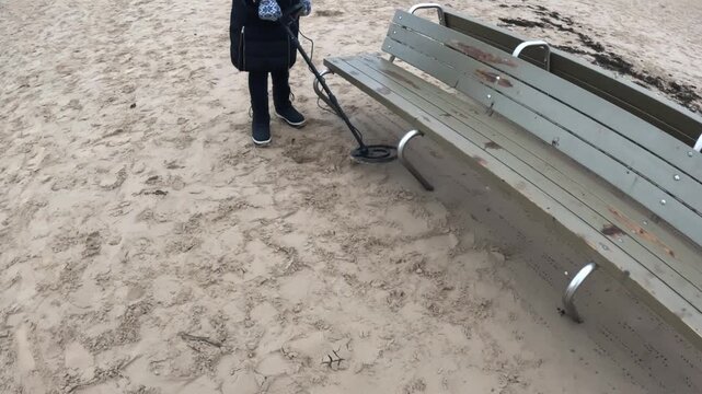Person scanning the sand around a weathered beach bench with a metal detector, searching for dropped items near a seating area at the coast.