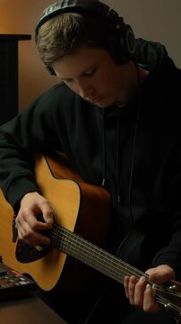 Music producer playing ascoustic guitar while composing track in dark home studio setting, vertical wearing headphones