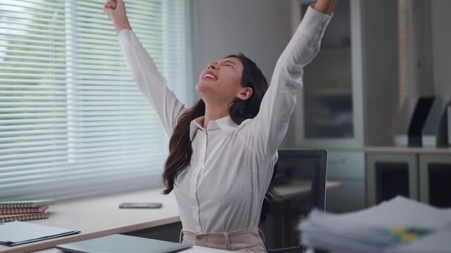 Happy asian business woman closing her laptop and stretching with arms raised after finishing a successful project in her modern office