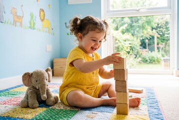 Happy toddler girl in yellow outfit stacking wooden blocks on a colorful mat in a sunny nursery room