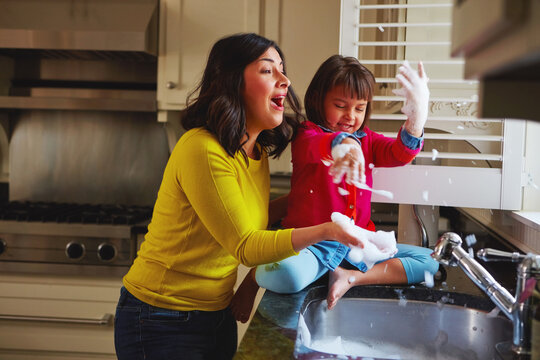 Mom, girl and play with soap, kitchen sink and happy with laugh, love and bonding at house. People, mother and daughter with games, bubbles or washing dishes with smile for learning in family home - Powered by Adobe