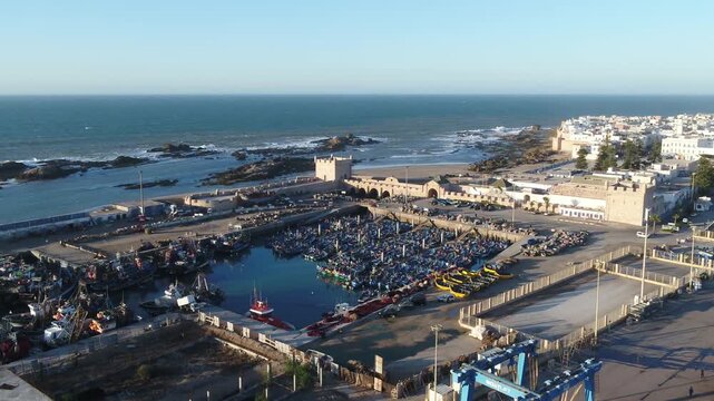Wide drone shot of Essaouira's fishing port featuring hundreds of blue boats against the backdrop of the vast Atlantic Ocean. Scenic coastal view.