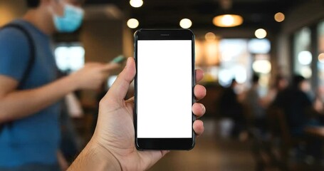 Person holding a smartphone with a blank white screen in a blurred cafe setting. - Powered by Adobe