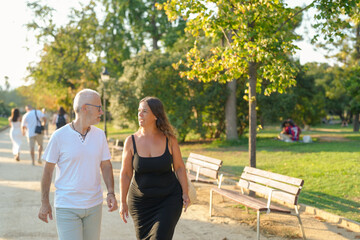Fototapeta premium A man and a woman are walking together in a park. The man is wearing a white shirt and the woman is wearing a black dress. They are walking on a path surrounded by trees and benches