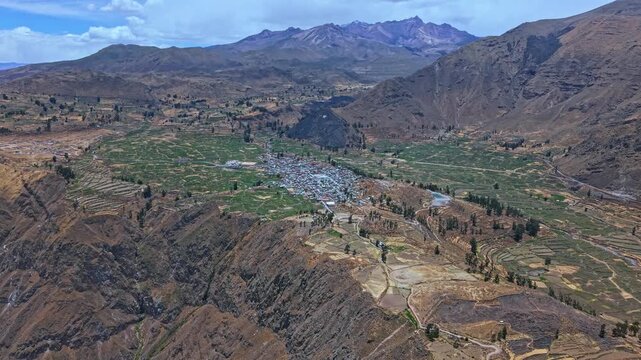 Epic drone footage captures Cabanaconde perched near the Colca Canyon edge. Terraced farmland and colorful Andean mountains unfold in a vast dramatic landscape showcasing scale geometry rugged beauty.