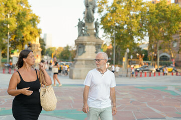 A man and a woman are walking down a street in a city. The woman is carrying a handbag and the man is wearing a white shirt. The scene is lively and bustling with people walking around