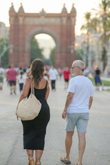 A woman and a man are walking down a street, with a large archway in the background. The woman is carrying a straw basket, and the man is wearing shorts. The scene is lively and bustling