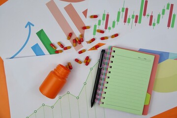 Vivid office scene showing a spilled orange pill bottle, capsules, a green notebook, a black pen, and assorted financial charts and colorful papers, suggesting health, study, and data analysis themes.