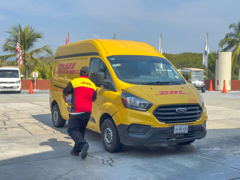 Huatulco, Oaxaca, Mexico. Dec 28, 2025. A delivery driver in uniform carries a parcel from a yellow DHL van parked in a sunny lot with palm trees and flags in the background.