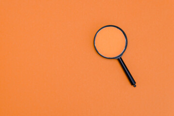 A black magnifying glass rests on an orange background. The glass is circular with a black handle, emphasizing clarity and focus.