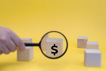 A hand holds a magnifying glass over wooden blocks with a dollar sign on one block. The background is bright yellow, emphasizing the financial theme.
