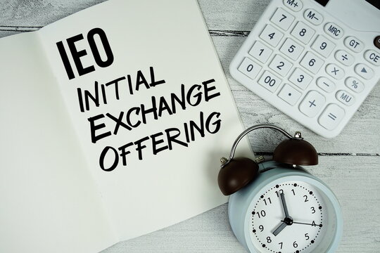 IEO Initial Exchange Offering text on paper notebook page with alarm clock and calculator on wooden background - Powered by Adobe