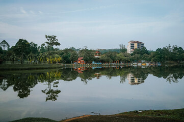 Fototapeta premium Serene lake reflection with lush green trees and buildings under a soft blue sky