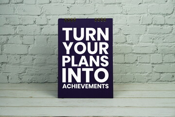 Fototapeta premium Turn Your Plans Into Achievements text on white brick wall and wooden background
