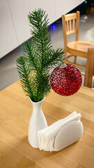 A red glitter Christmas ball on a spruce branch in a white vase on a wooden table in a cafe.