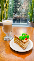 A slice of layer honey cake with green cream decoration and a glass of latte on a wooden cafe table.