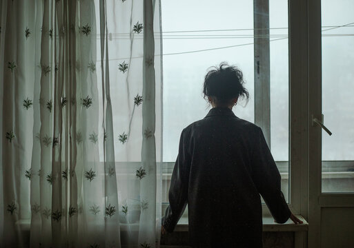 An elderly woman stands at the window in winter