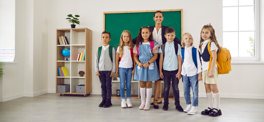 Teacher and students in classroom during learning. A smiling teacher stands with diverse group of children wearing backpacks by the chalkboard, ready for class. Ideal back to school concept.