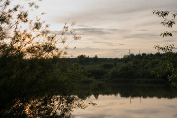 A tranquil river scene at sunset, surrounded by lush greenery and trees. The water reflects the soft colors of the sky, creating a peaceful atmosphere.