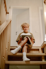 A child with curly hair sits comfortably on wooden stairs, wearing headphones and focused on a...