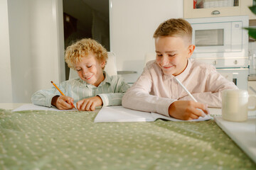Two children are seated at a table, focusing on their school assignments. They are writing in notebooks, surrounded by a clean and bright kitchen.