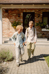 Two young boys with backpacks are walking along a path in front of a house. They are smiling and chatting, enjoying a sunny afternoon after school.