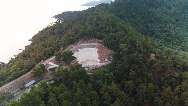 Aerial drone circle shot around the ancient Greek Theater of Limenas, Thasos Island, highlighting the historic ruins and surrounding coastal town at golden hour sunrise.