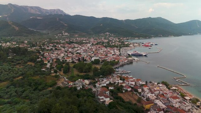 Wide aerial drone establishing shot panning over Limenas town, the capital of Thasos Island, Greece, revealing the harbor, coastal architecture, and mountains at golden summer sunrise.