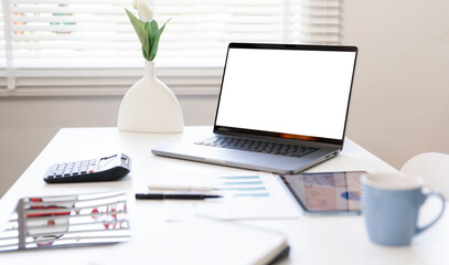 Laptop mockup with blank white screen on a bright office desk with a coffee cup, calculator, and vase. Minimalist workspace with ample copy space.