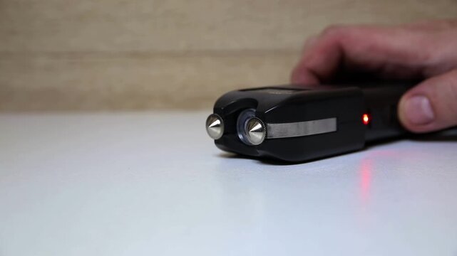 Close-up view of person hand holding black stun gun. Device rests on white surface, wooden wall in background. Bright electric discharge flashes between metal electrodes of self-defense weapon.