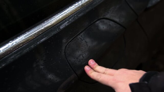 Close up detail of hand of person pressing car body to open fuel tank cover. Black vehicle exterior shows water drops from recent rain. Preparation for gas fill up.