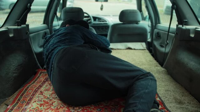 Person rests in back of car, vehicle converted for sleeping accommodation. Background shows front seats of automobile. Scene depicts life on road, struggle of homelessness