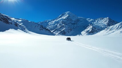 Black SUV drives across a vast white snow field towards high mountain peaks under a blue sky. A scene of adventure and winter exploration suitable for travel and automotive concepts.