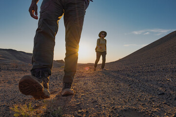A couple walks a dirt road in the Mongolian desert towards the camera at sunset. Their long shadows and boot soles are visible as they move against the setting sun.