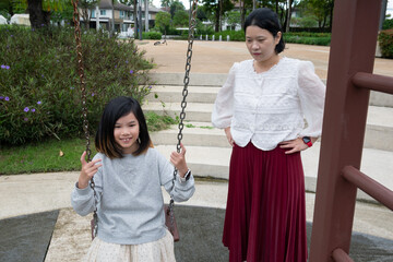 Happy Asian mother gently pushing her daughter on a swing in the outdoor playground, enjoying active family bonding time