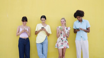 Group of multicultural young friends standing against a yellow wall, smiling and absorbed in their mobile phones, typing and chatting online - Powered by Adobe