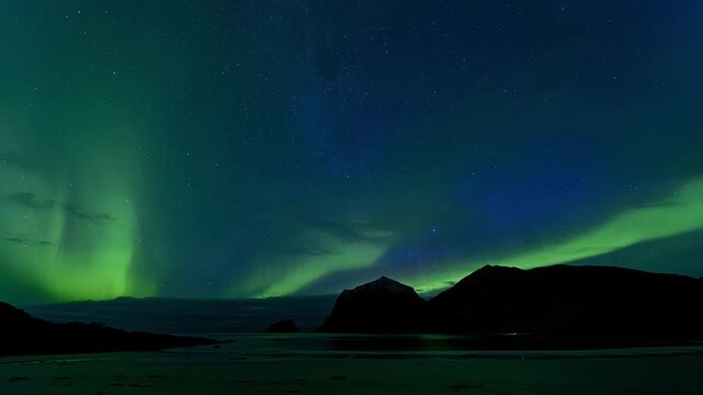 Starry skay and aurora borealis northern lights over sandy beach surranded by mountains by the sea; flashing lighthouse far in the distance. Lofoten Islands, Northern Norway. Time lapse. 4K