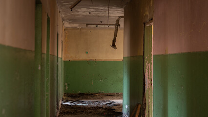 In an abandoned building, a long corridor shows peeling walls with a green lower half. Broken doors lead to empty rooms and debris covers the dusty floor. Light comes from the end.