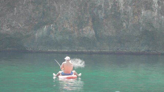 Man fishing, sea, raft, smoking e-cigarette on a sunny day in clear water near a rocky cliff