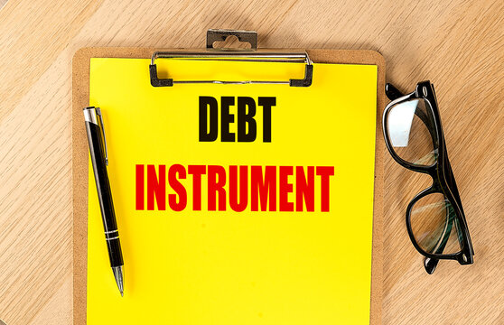 Clipboard with text debt instrument on yellow paper with pen and eyeglasses on wooden desk representing financial tools