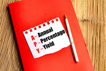 Acronym APY with the words annual percentage yield written on torn paper on red notebook with pen on wooden table